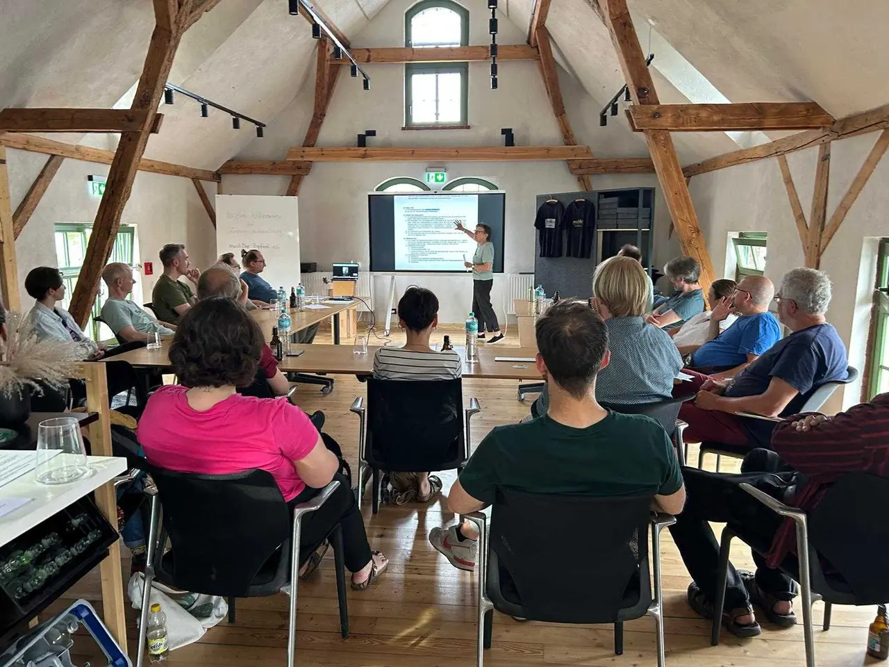 Gründungsversammlung der machBar Zapferei eG im Kranhaus: rund zwanzig Menschen sitzen unter dem offenen Holzdachstuhl und verfolgen eine Präsentation.